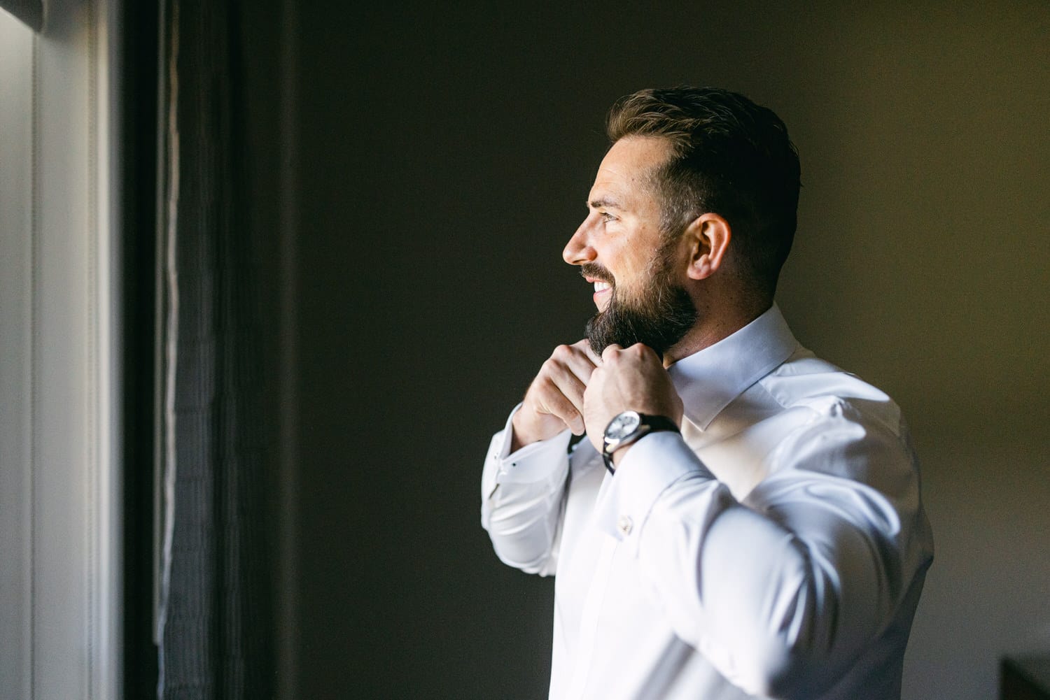 A smiling man in a white shirt adjusts his collar while standing by a window, showcasing a well-groomed beard and a stylish watch.