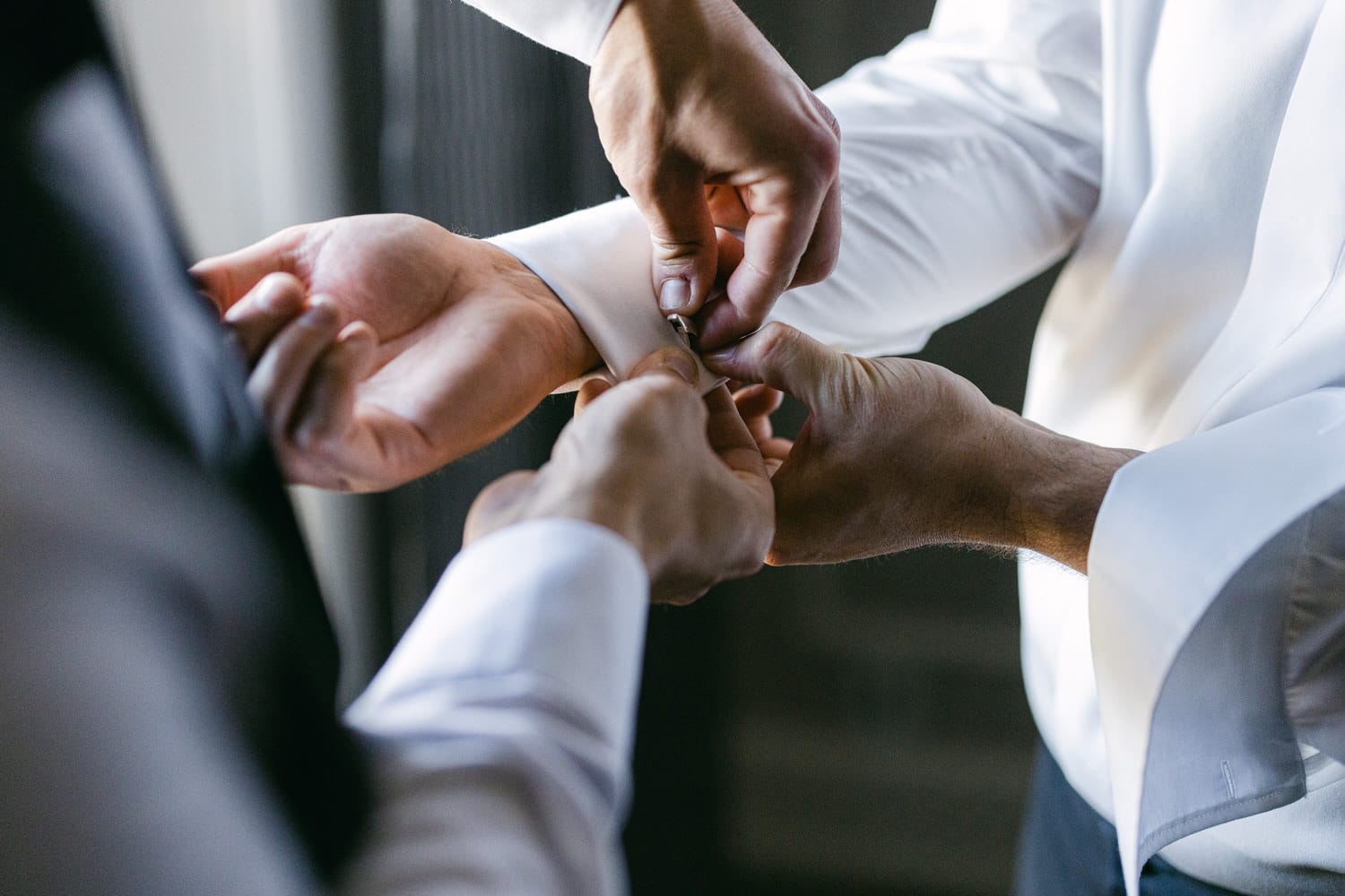 Two people assist in fastening a cufflink on a formal shirt in a well-lit setting.