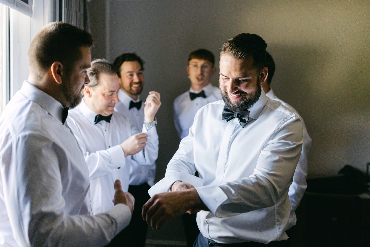 A group of six men in white dress shirts and black bow ties getting ready, with one man adjusting his cuff while others help each other with attire in a well-lit room.