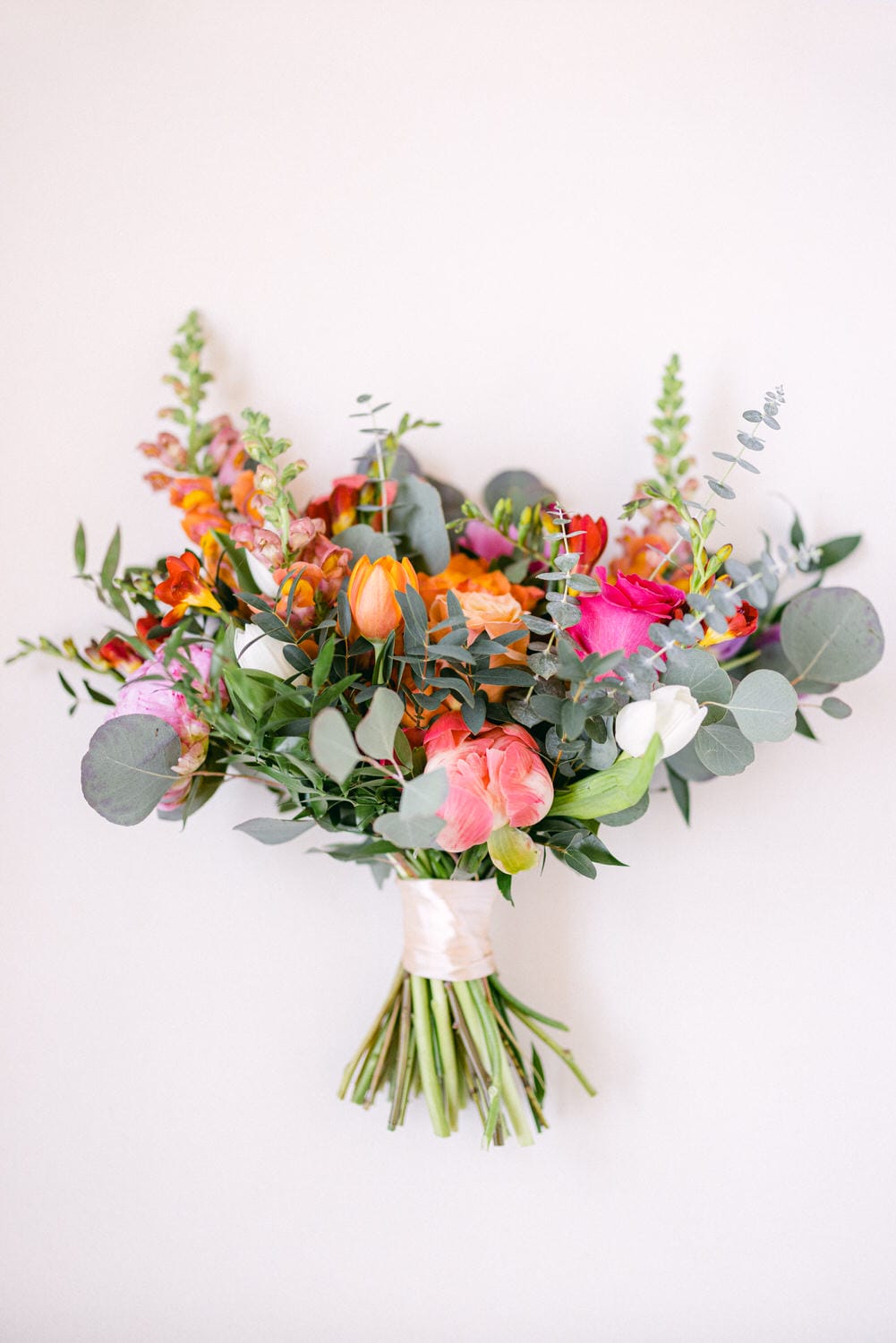 A colorful bouquet featuring a mix of pink, orange, and white flowers, elegantly wrapped with ribbon and surrounded by greenery, set against a neutral background.