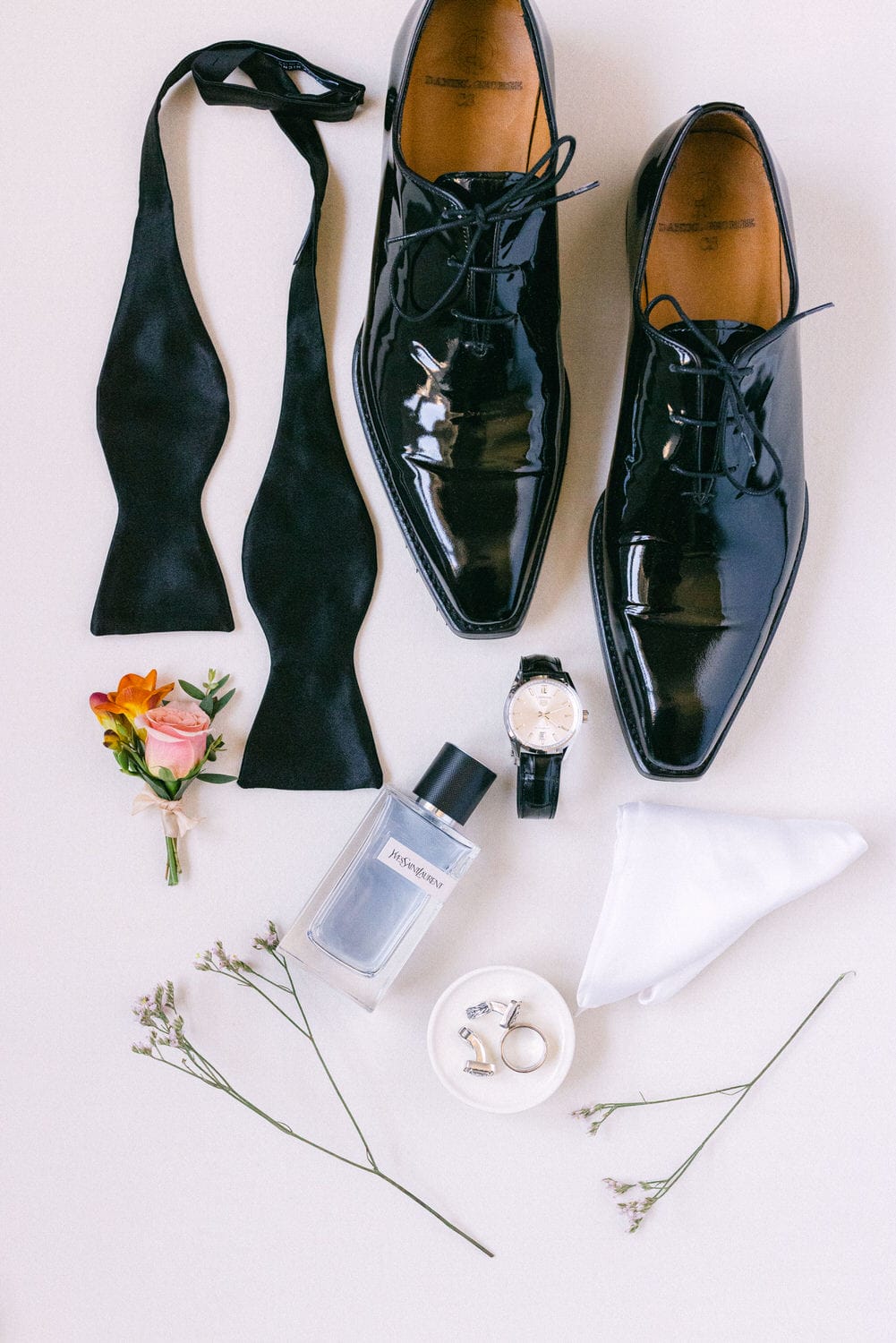 A flat lay of polished black dress shoes, two black bow ties, a silver watch, a bottle of perfume, a flower bouquet, a white pocket square, and elegant rings against a light background.