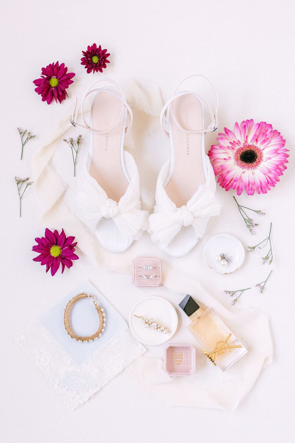 A flat lay capturing stylish white shoes adorned with bows, surrounded by vibrant flowers, jewelry, a delicate perfume bottle, and decorative fabric.