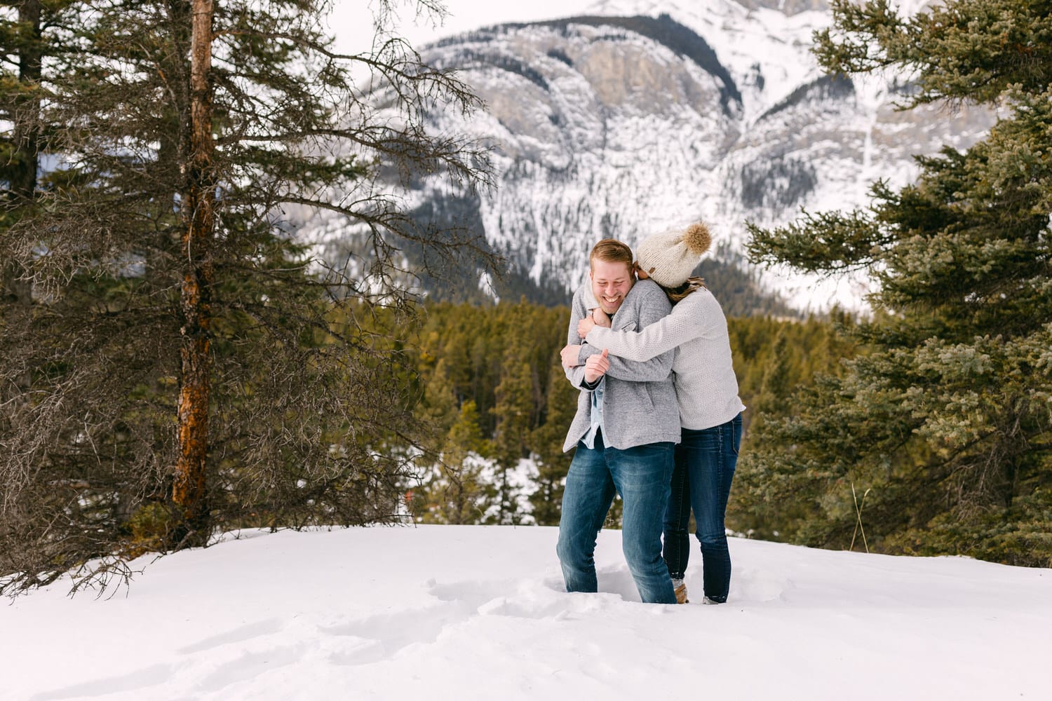 A couple embraces playfully in the snow-covered landscape, surrounded by towering pine trees and majestic mountains in the background.