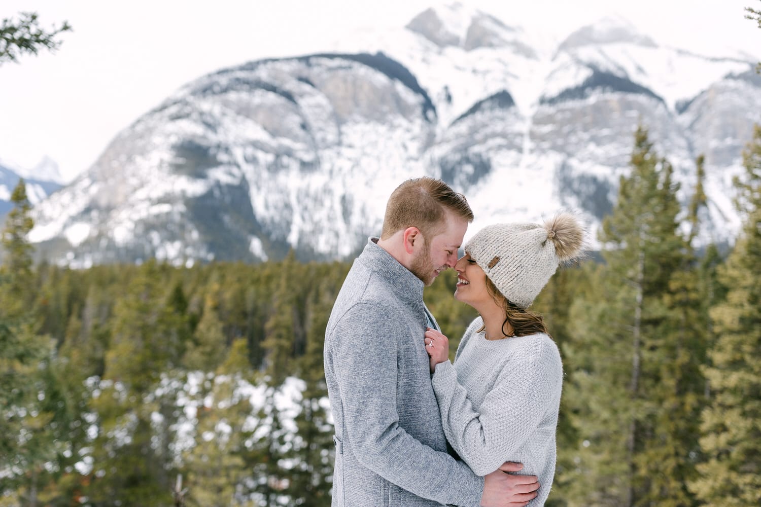 A couple shares a joyful moment, forehead to forehead, surrounded by snowy mountains and evergreen trees.