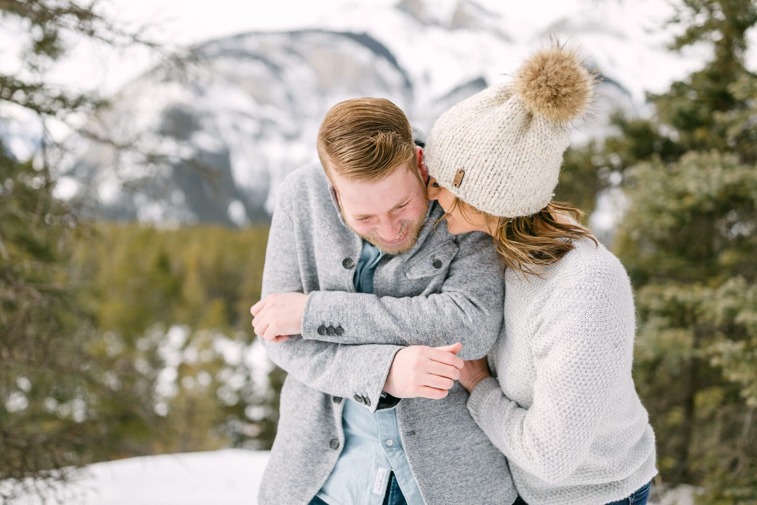 A couple shares a joyful moment while embracing in a snowy mountain landscape, dressed warmly in cozy winter attire.