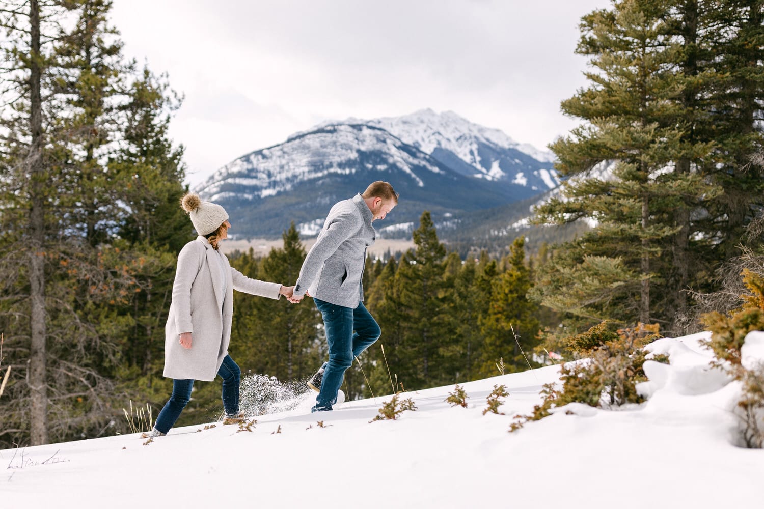 A couple holding hands while walking through a snowy landscape with pine trees and mountains in the background.