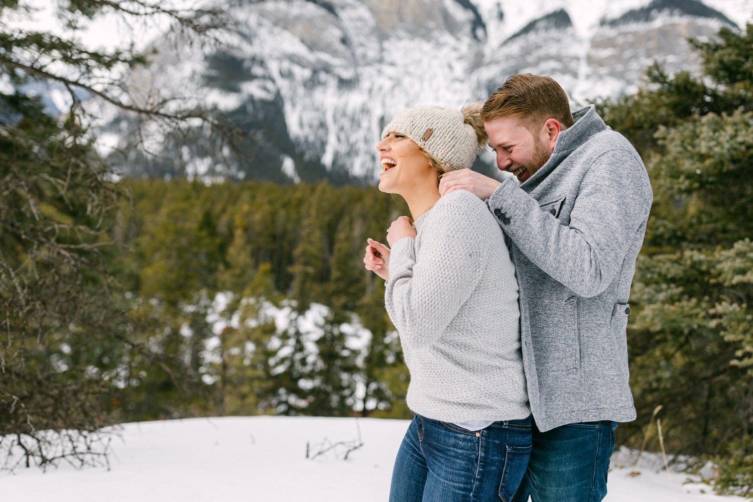 A couple shares a laugh while enjoying a snowy landscape, surrounded by mountains and evergreen trees.