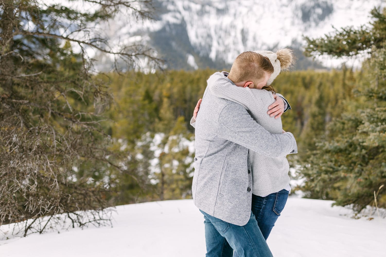 A couple warmly hugging in a snowy landscape surrounded by evergreen trees and mountains.