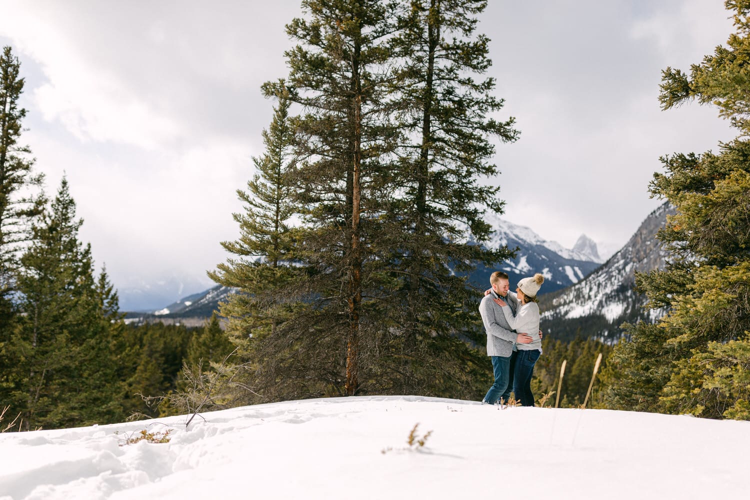 A couple embracing in a snowy landscape, surrounded by evergreen trees and towering mountains in the background.