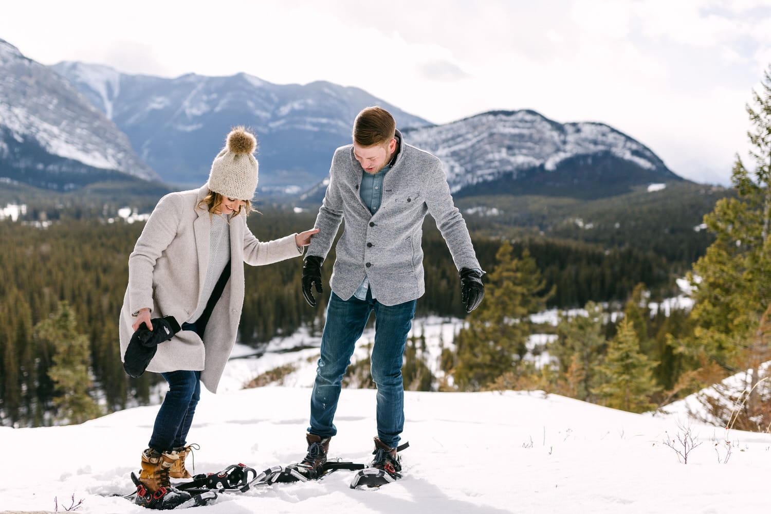 A couple enjoys snowshoeing in a snowy mountain landscape, with trees and peaks in the background.