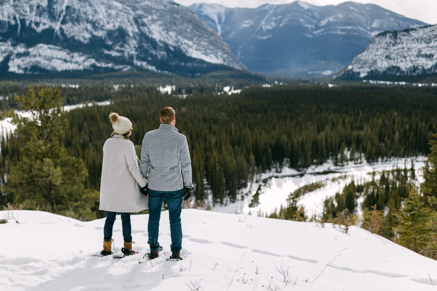 A couple stands hand in hand on a snowy hillside, overlooking a verdant valley and majestic mountains in the distance.