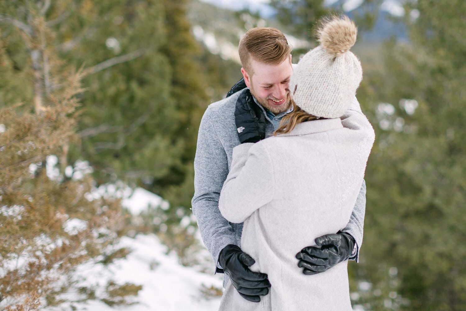 A loving couple shares a warm embrace in a snowy forest, surrounded by evergreen trees.