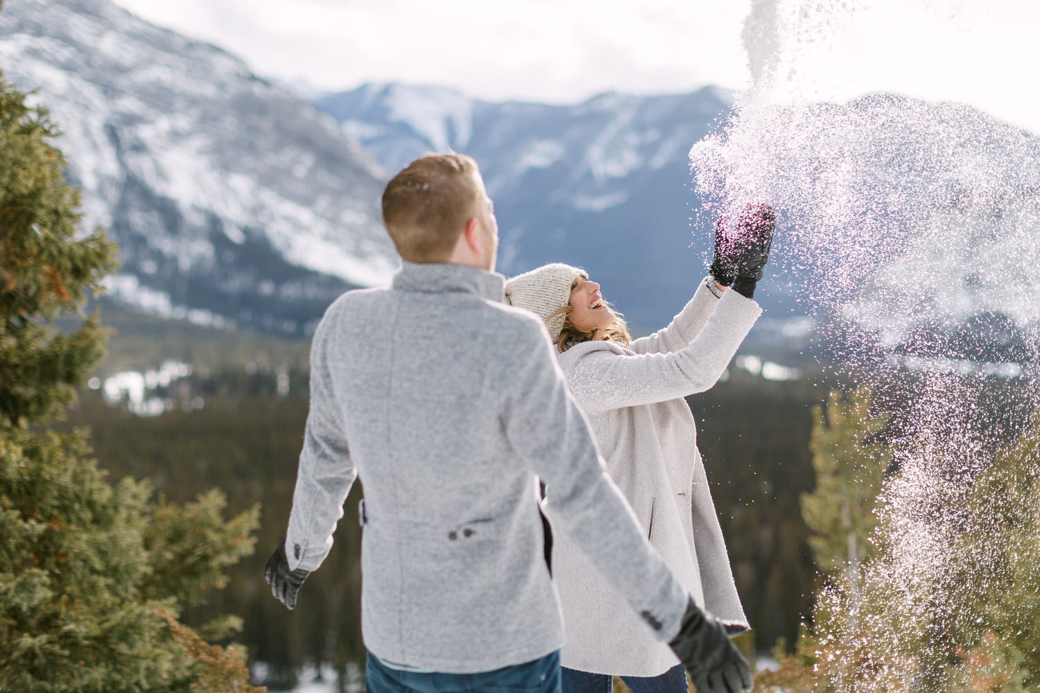 A couple joyfully playing in the snow-covered mountains, with one throwing snowflakes into the air while the other watches.