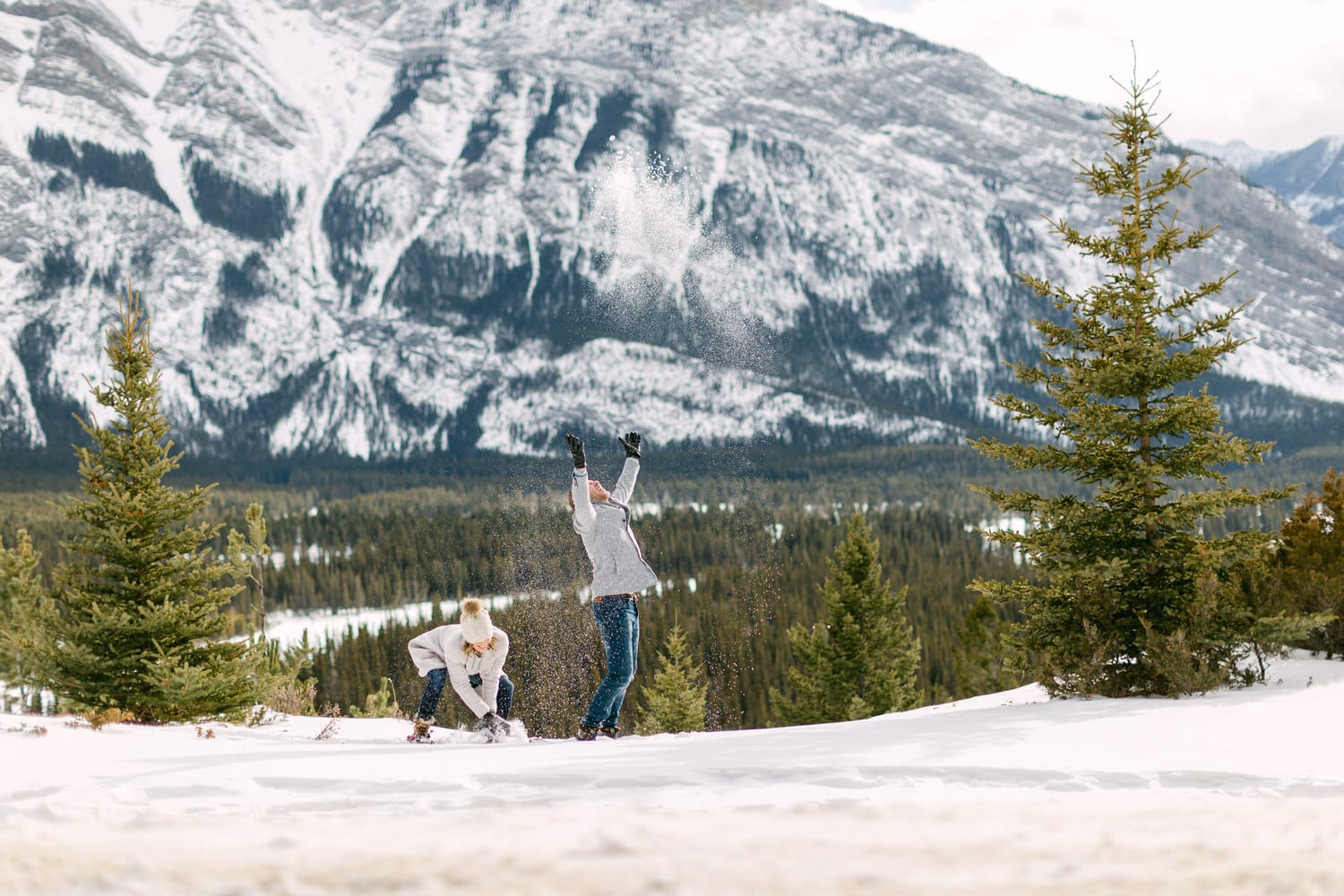 Two people playing in the snow with a stunning mountain backdrop, one throwing snow into the air and the other crouched down.