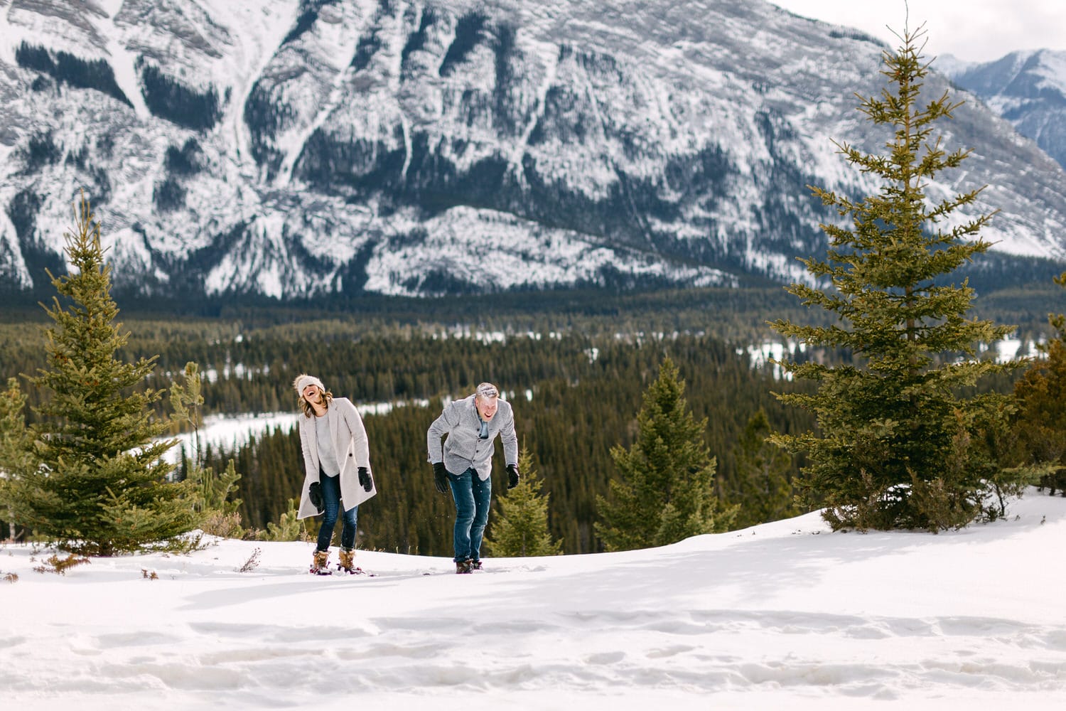 Couple smiling and snowshoeing in a snowy landscape with mountains and evergreen trees in the background.