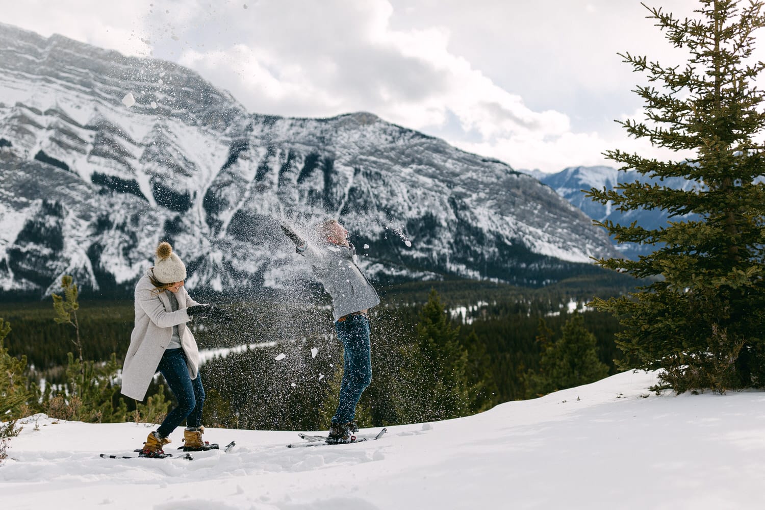 Two people playing in the snow, throwing snow at each other against a backdrop of snowy mountains and evergreen trees.