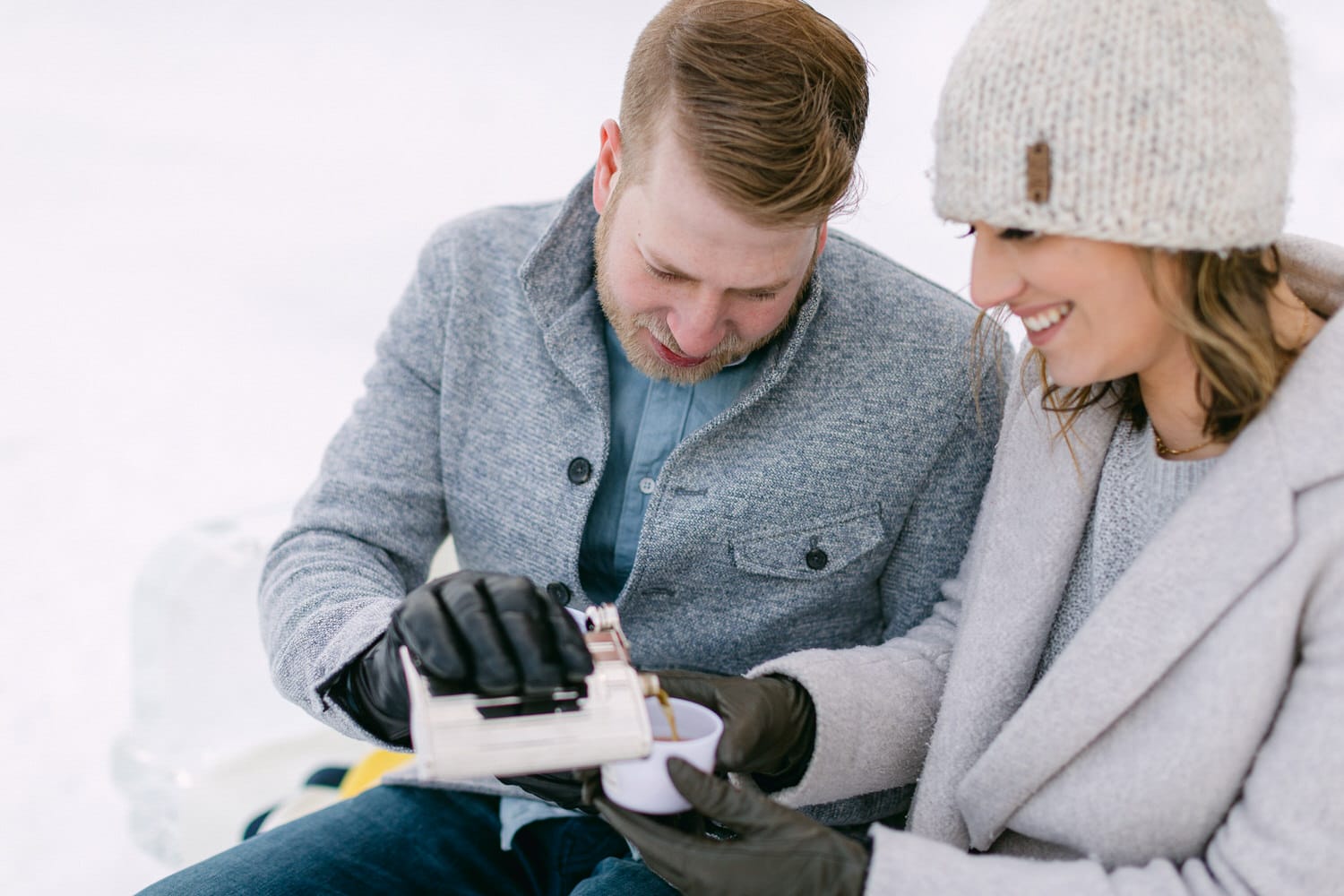 Cozy Winter Moments::A couple joyfully sharing a warm drink while seated outdoors in the snow, dressed in winter attire.