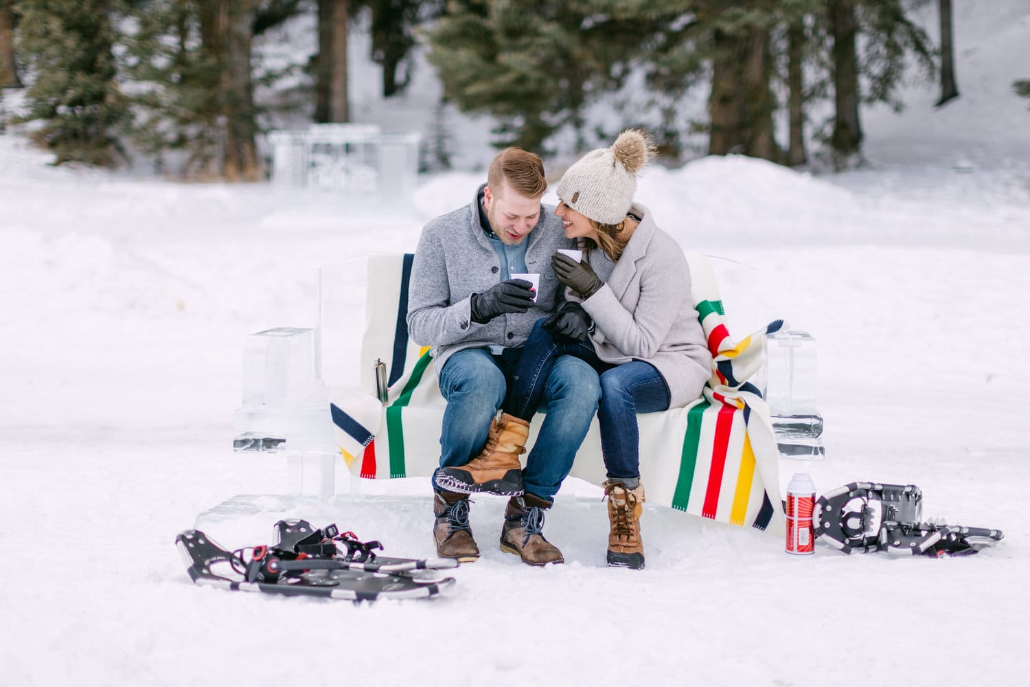 Couple enjoying a snowy day together on an ice bench, sharing a warm drink and laughter amidst winter scenery.