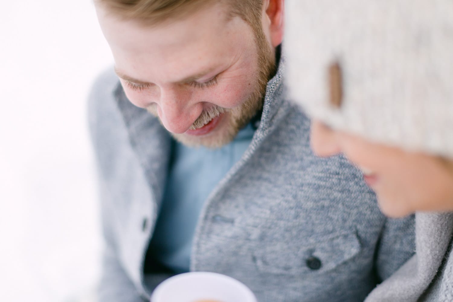 A close-up of a smiling couple sharing a light-hearted moment, with one holding a cup, dressed warmly against a snowy backdrop.