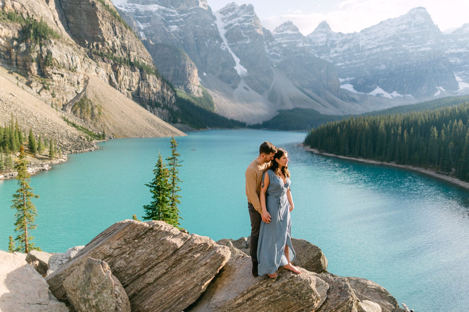 A couple stands on a rocky outcrop near a vibrant turquoise lake, surrounded by mountains and evergreen trees, capturing a moment of intimacy and connection.