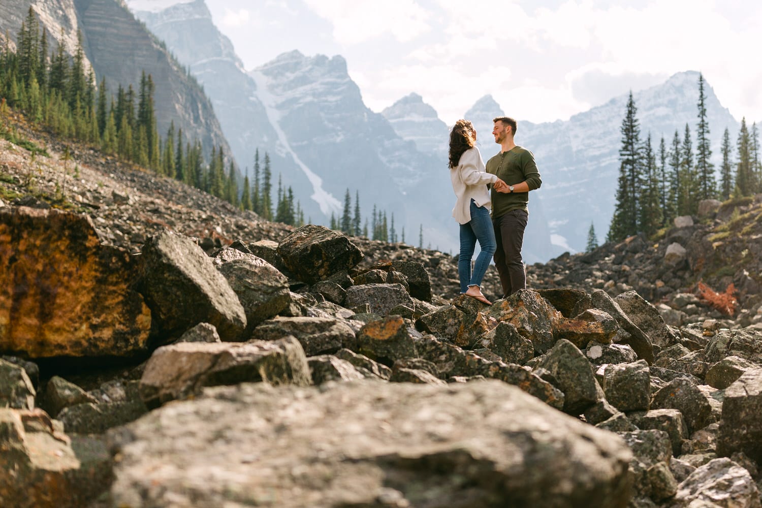 A couple stands on rocky terrain surrounded by majestic mountains, holding hands and enjoying a moment together.