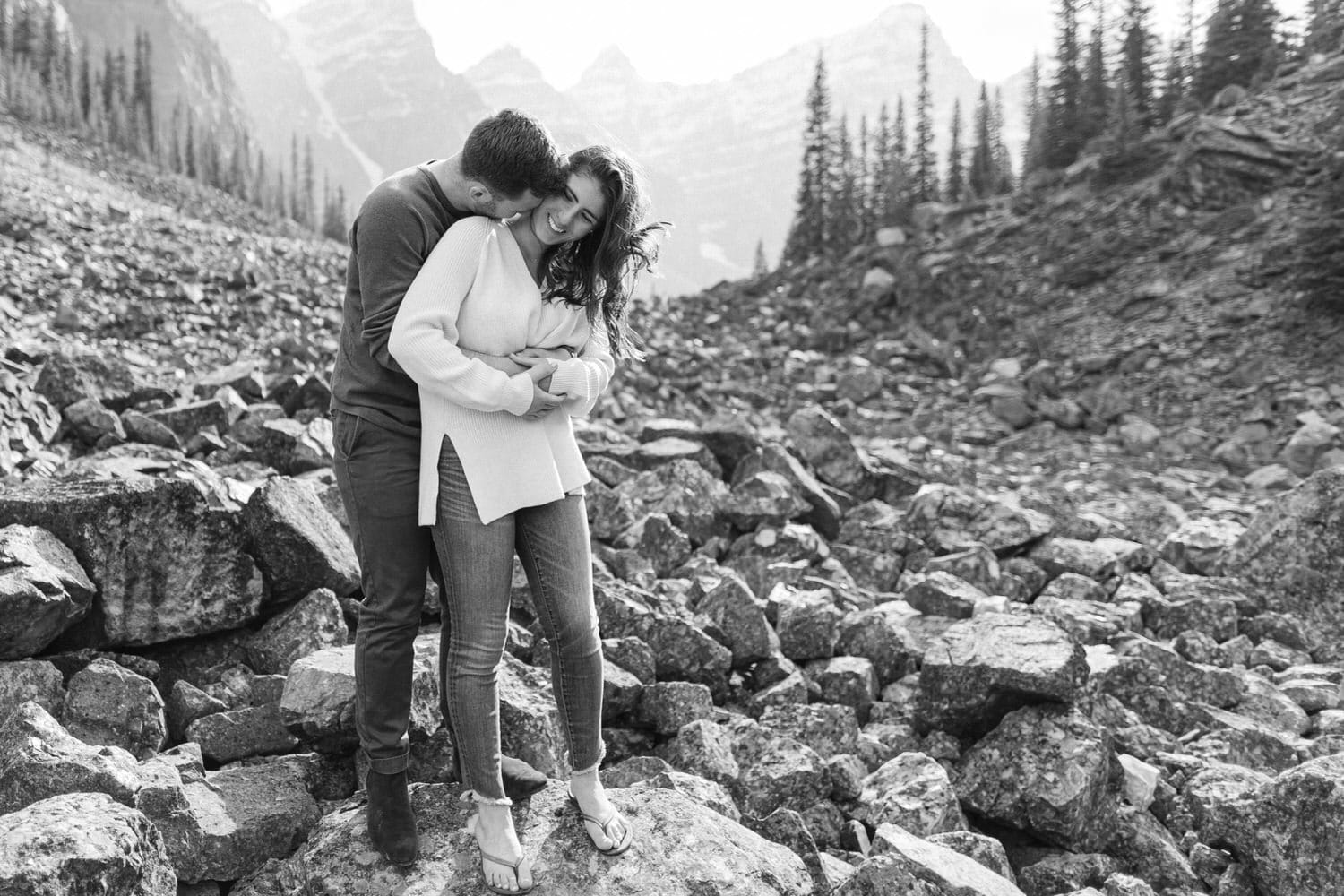 A joyful couple embraces outdoors among rocky terrain, surrounded by towering mountains and evergreen trees.