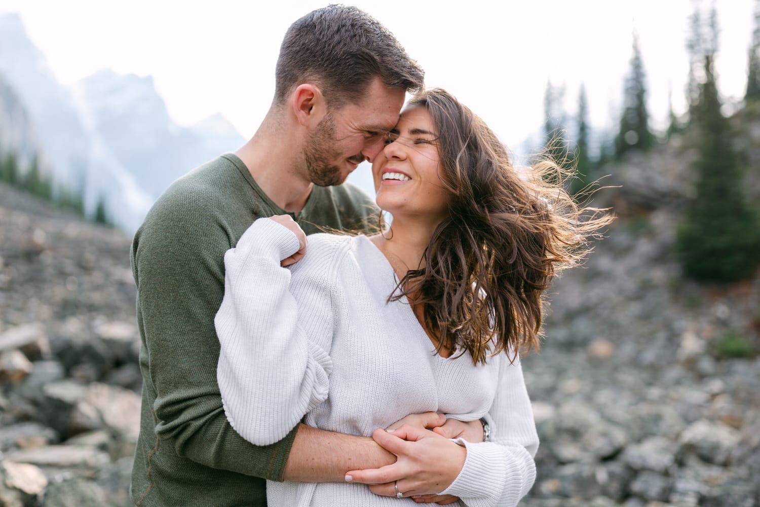 A joyful couple sharing a tender moment outdoors, surrounded by a rocky landscape and distant mountains, with the woman laughing and the man leaning in closely.