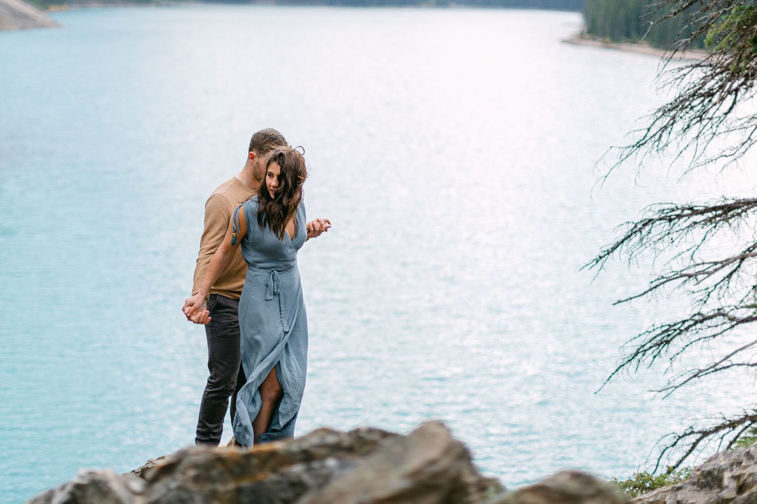 A couple stands on rocky terrain near a calm blue lake, holding hands and gazing at each other, with forested hills in the background.