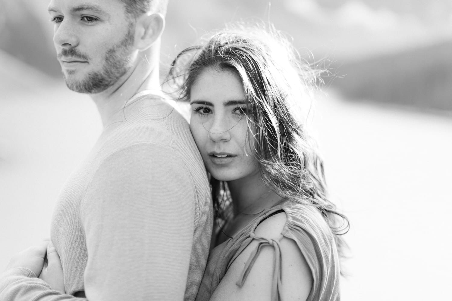 A black and white portrait of a couple; the woman gazes intently at the camera while the man, seen from the side, leans slightly backwards, both embodying a sense of intimacy and connection.