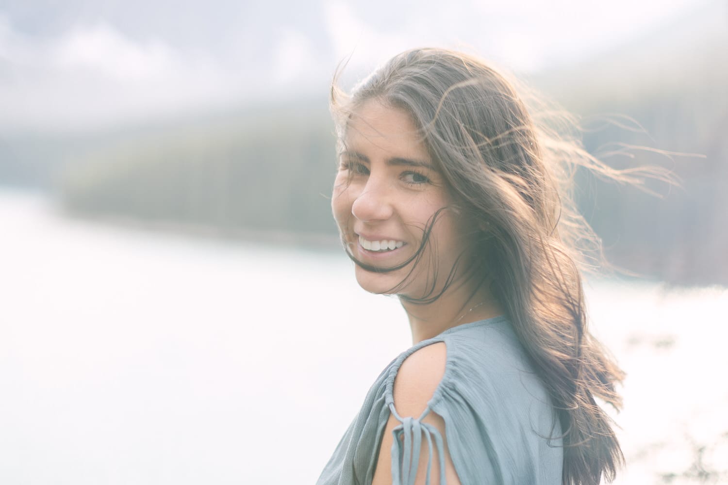 A woman with flowing hair smiles gently at the camera, with a serene lake and mountains in the background.