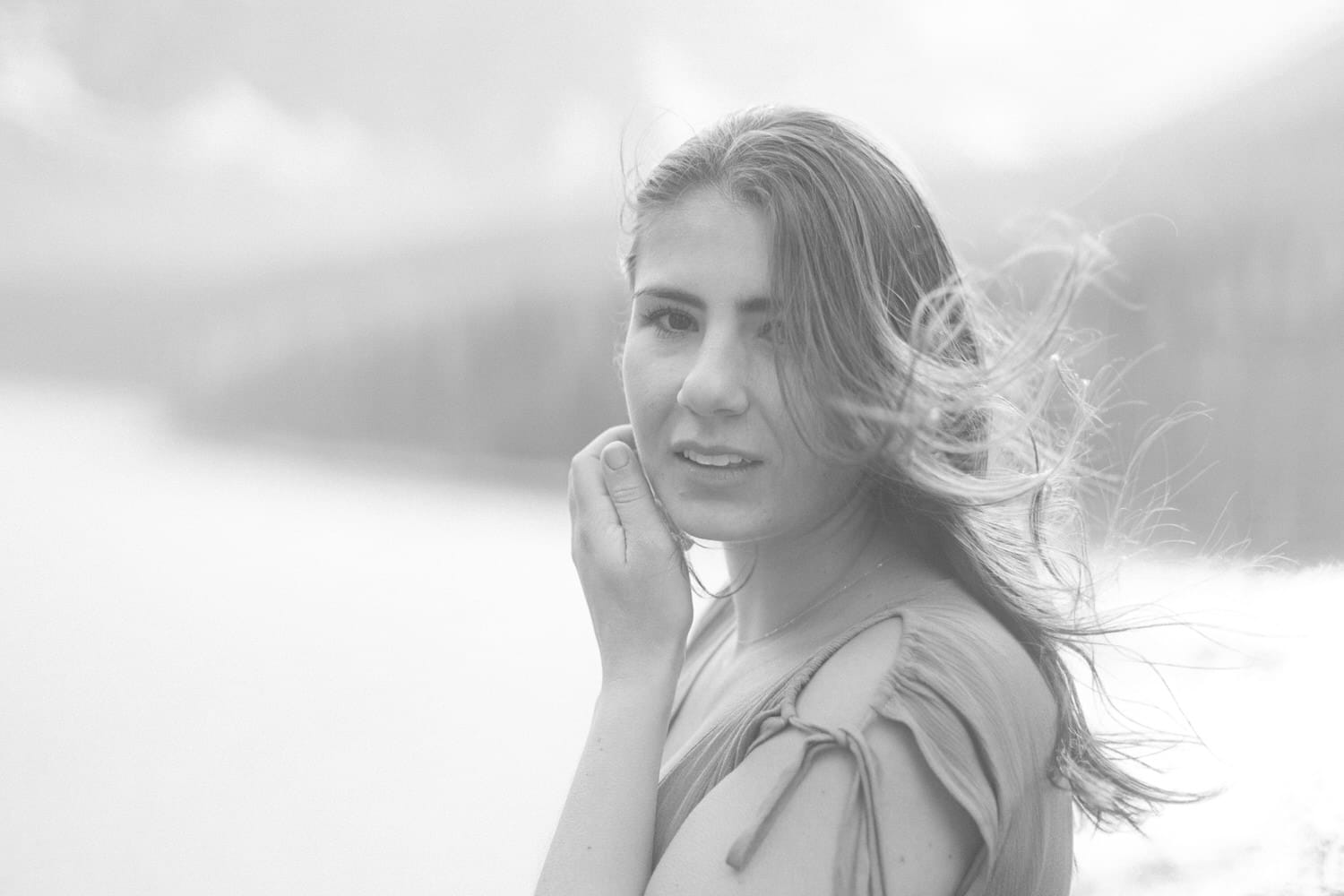 A woman with flowing hair looks directly at the camera, softly holding her face, surrounded by a misty landscape in black and white.