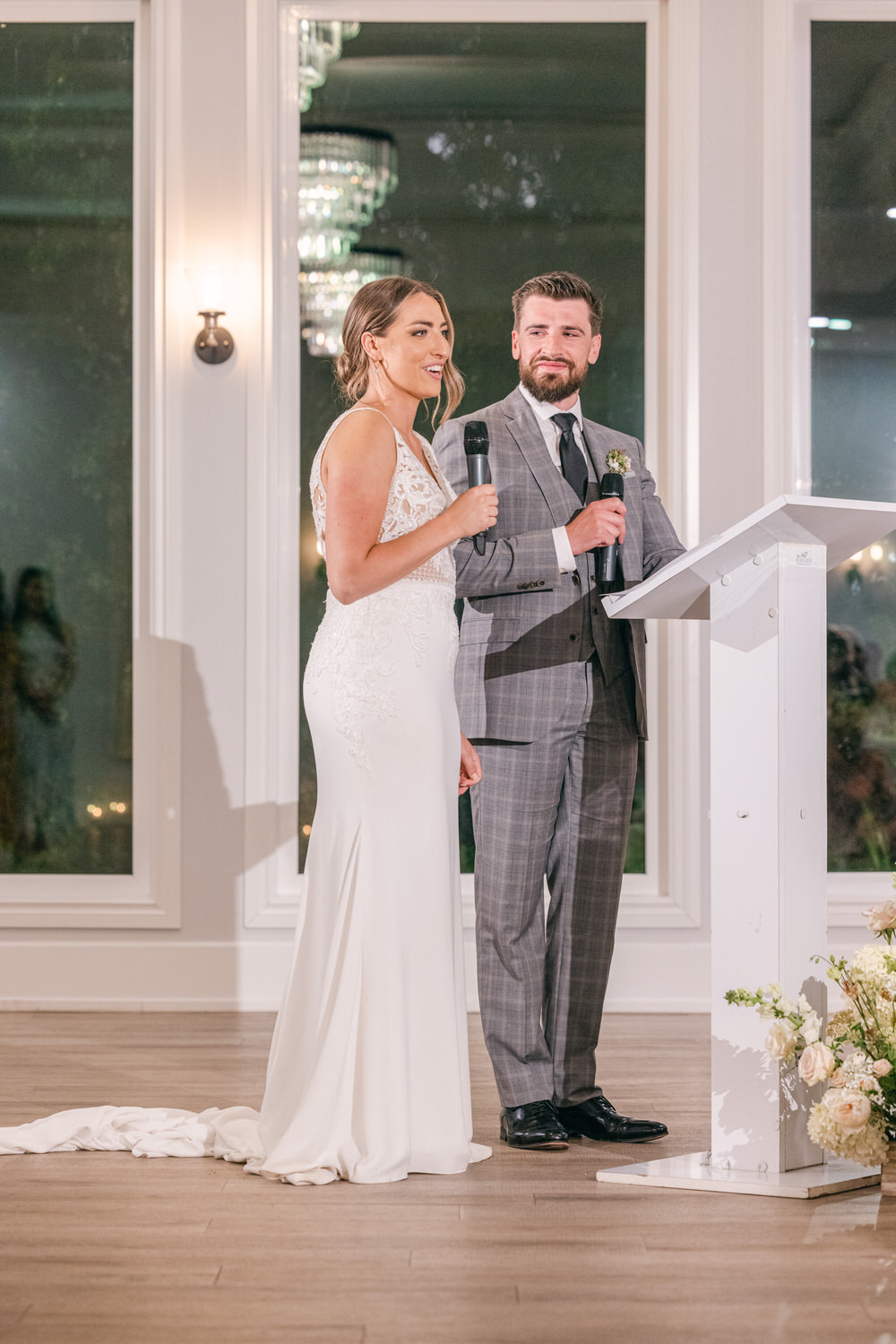 A bride in a white dress and a groom in a grey suit stand together, speaking at a wedding reception with elegant decor in the background.