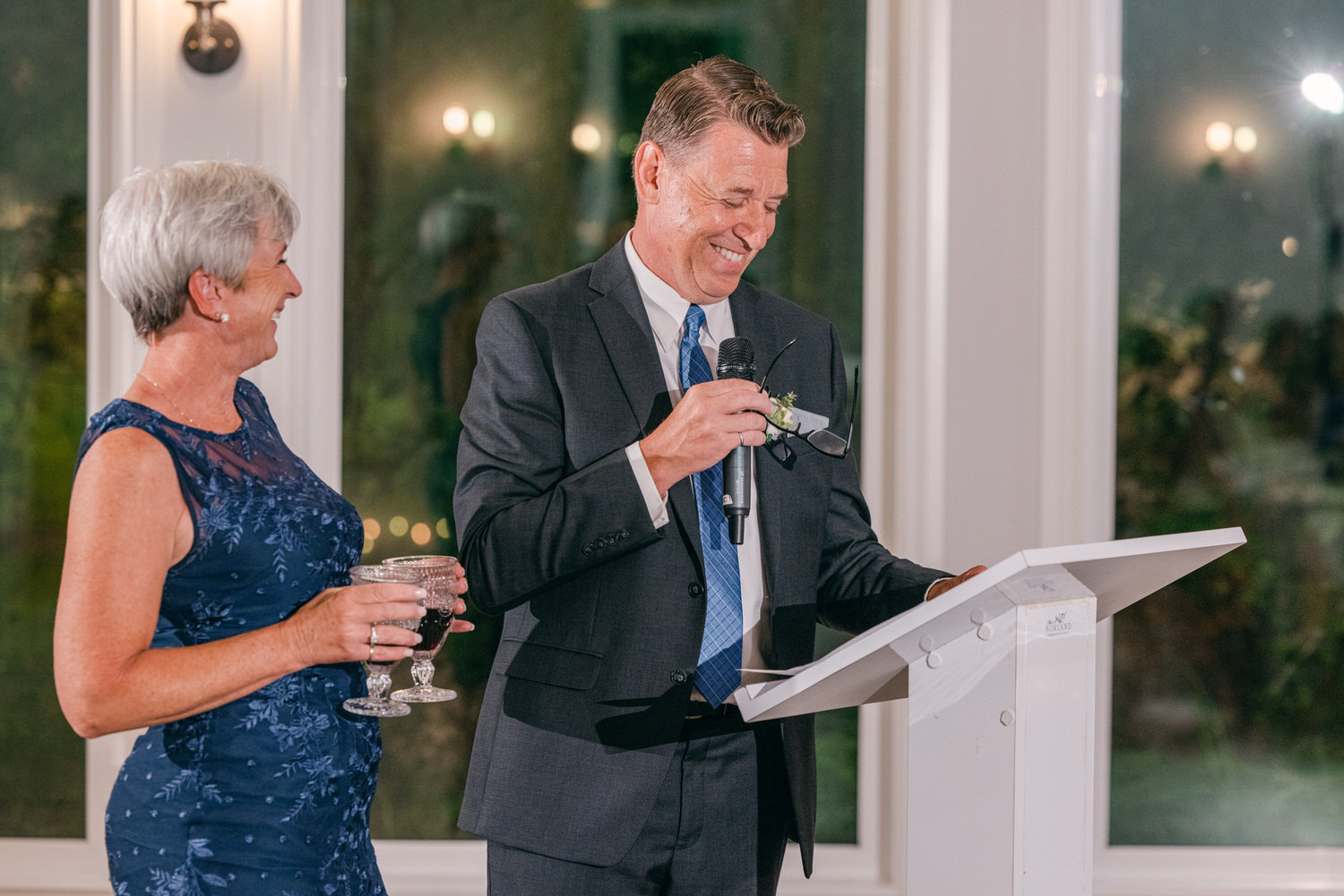 A man in a suit delivers a speech while a woman in a blue dress smiles, holding two glasses, at a wedding reception.