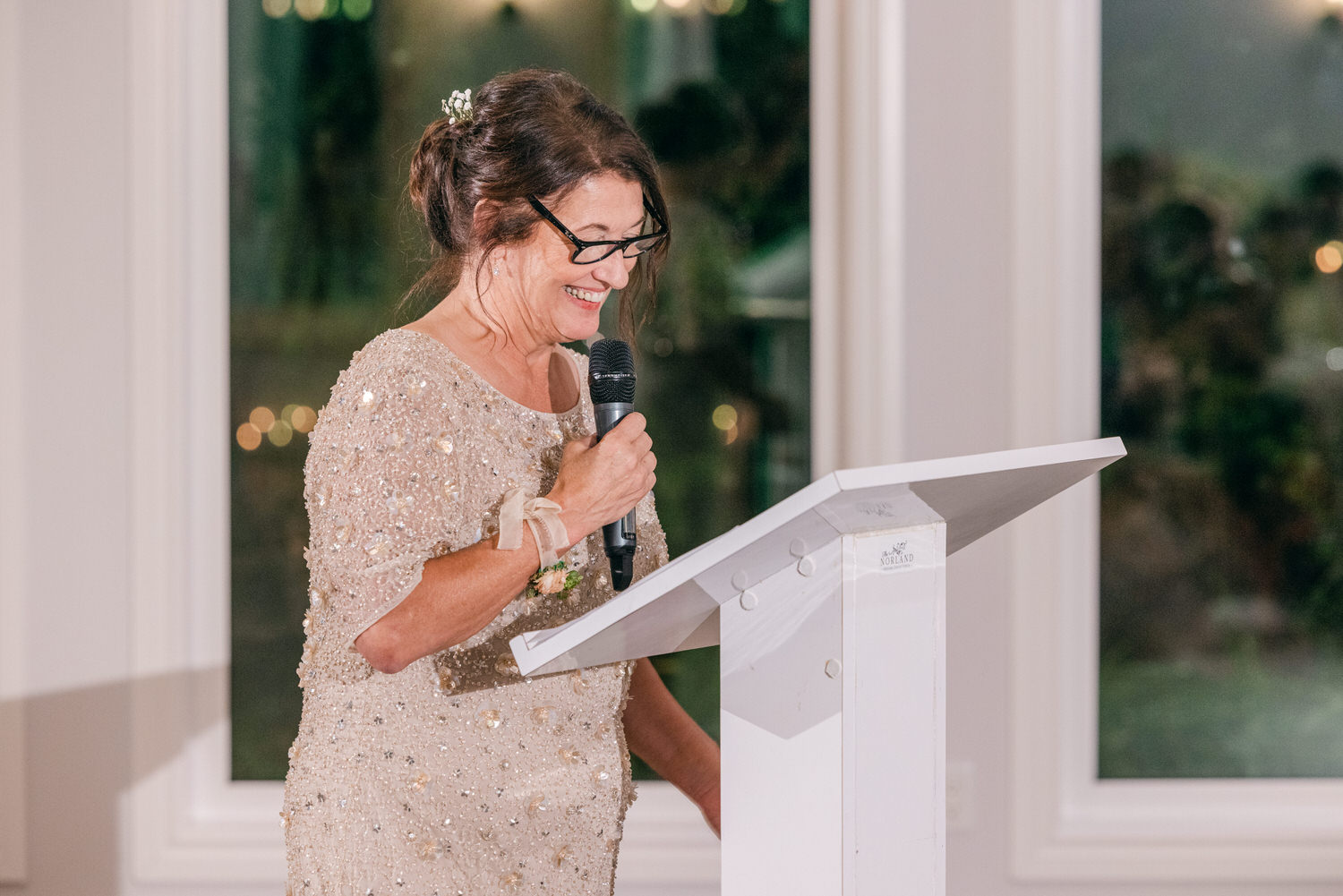 A woman in a sparkling dress delivers a heartfelt speech at a wedding reception, holding a microphone and smiling.