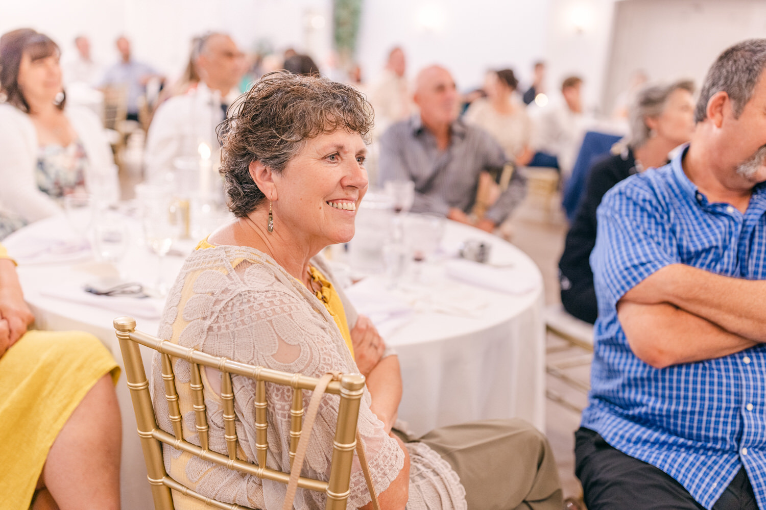 A woman with short, curly hair laughs while seated among an audience at a festive gathering, showcasing a joyful atmosphere.