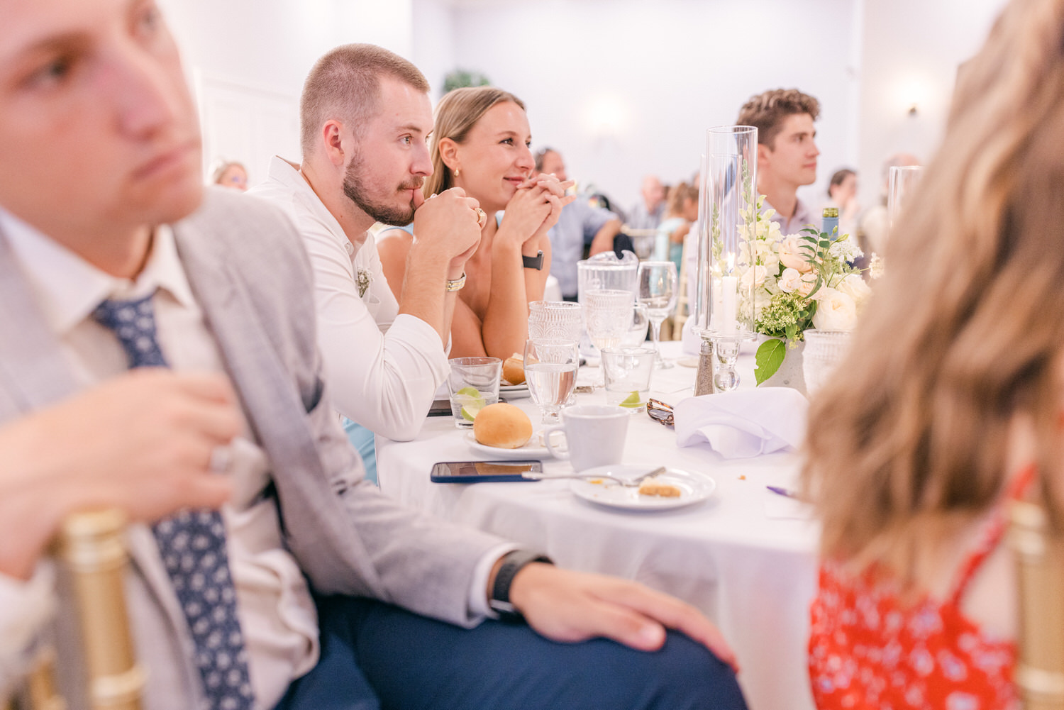 Guests attentively listening during a wedding reception, with elegant table settings and floral decorations in the background.