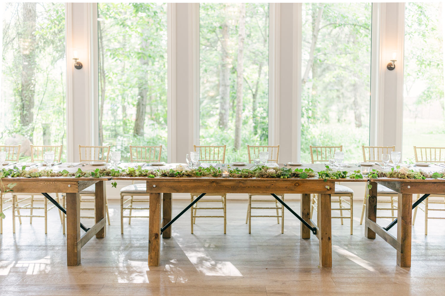 A beautifully arranged dining table with greenery and floral decorations, set for an event in a bright space surrounded by large windows and natural light.