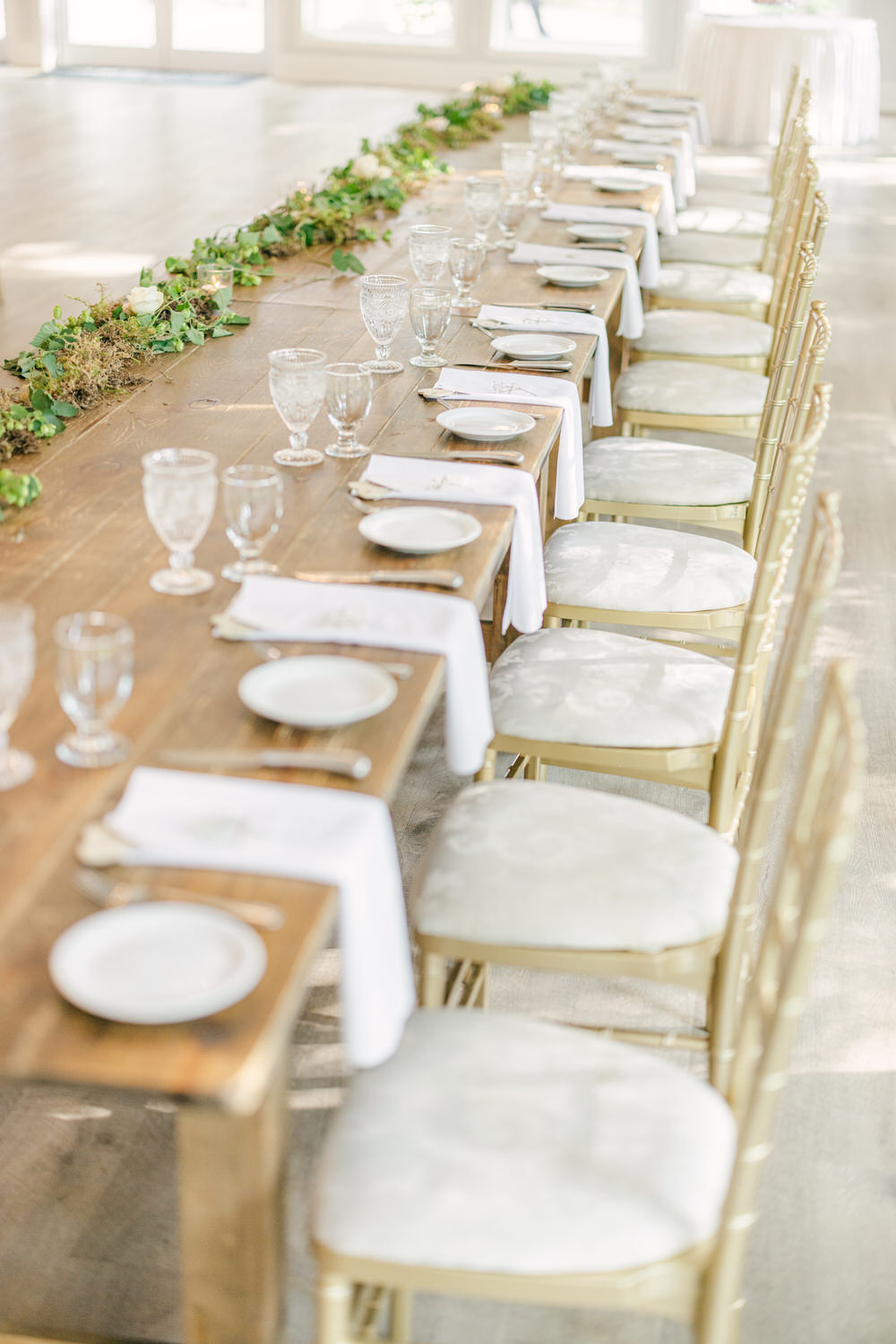 A long wooden dining table elegantly set with white napkins, delicate glassware, and decorative greenery, ready for a gathering.