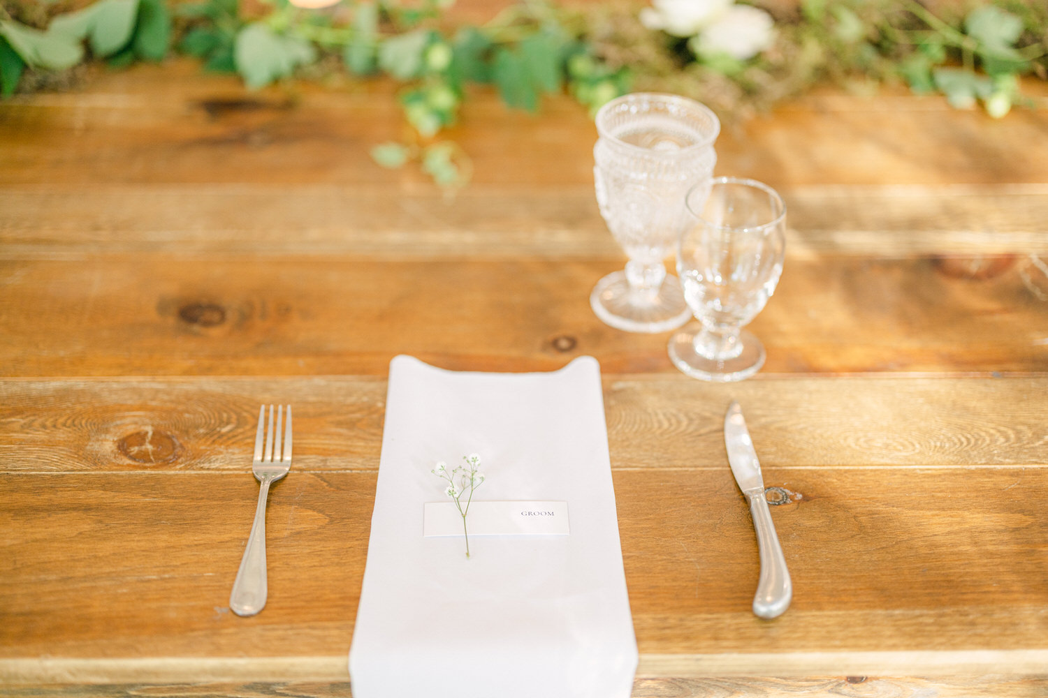 A neatly arranged table featuring a white napkin with a place card reading "GROOM," accompanied by a small flower, forks, knives, and elegant glassware on a polished wooden surface.