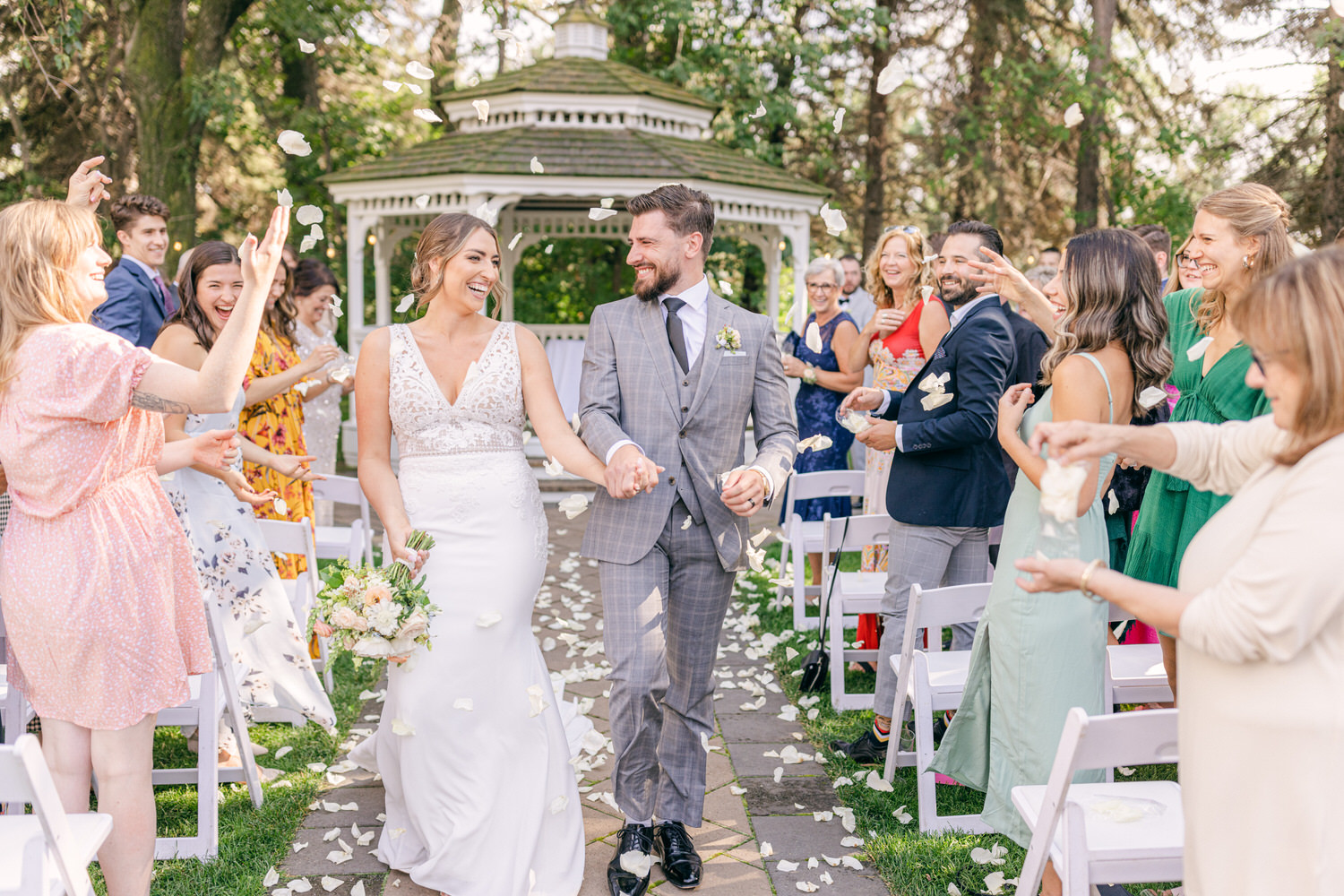 A newlywed couple walks hand in hand down an aisle, surrounded by smiling guests tossing flower petals in a garden setting.