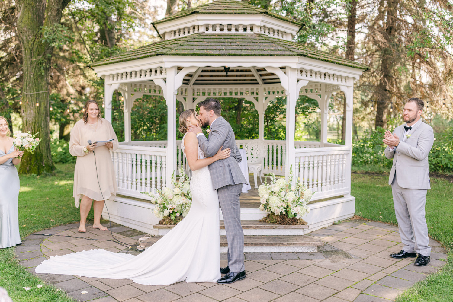 A couple shares a kiss during their wedding ceremony at a gazebo, surrounded by a wedding party and floral arrangements.