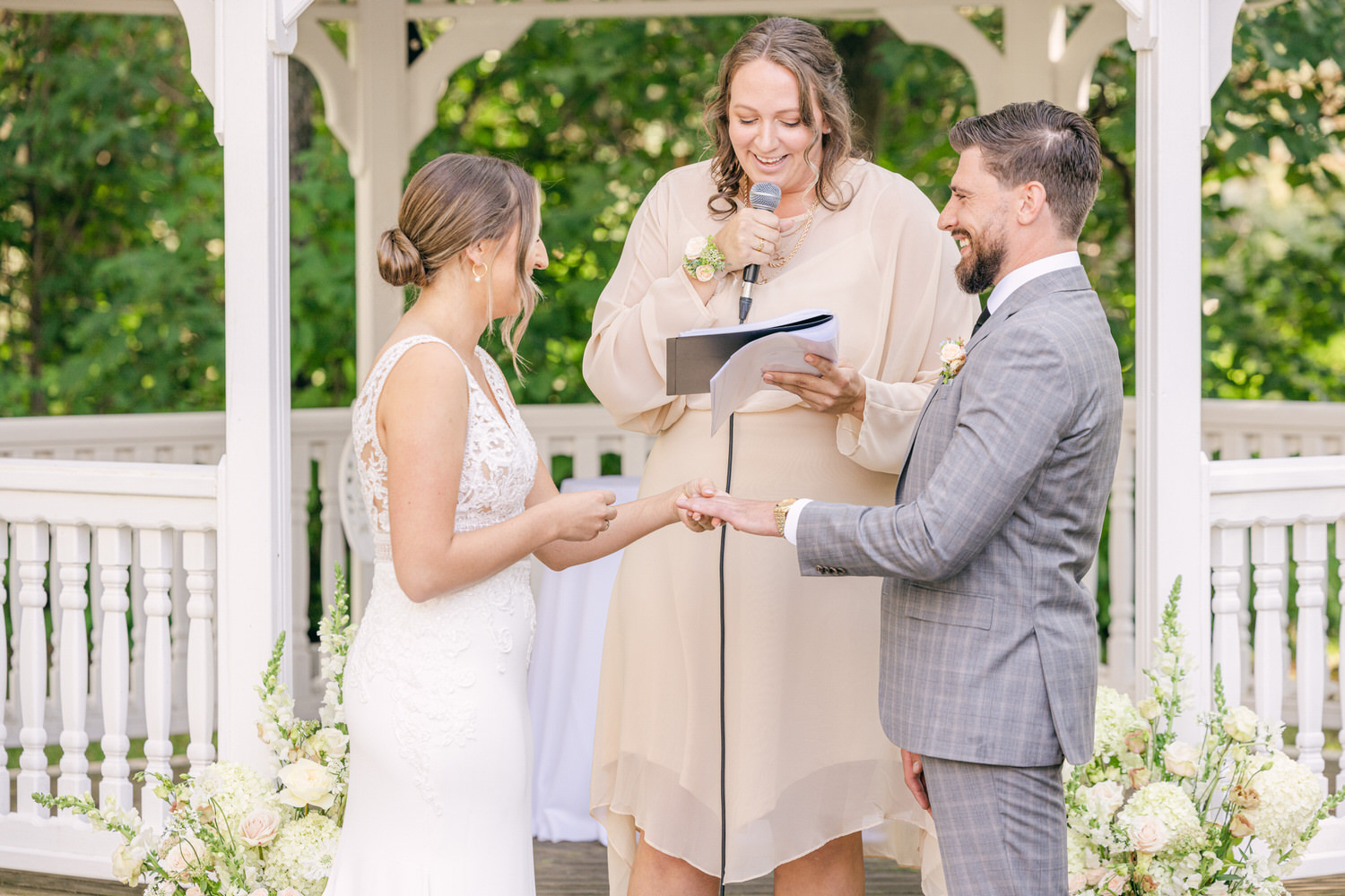 A bride and groom exchanging vows during their wedding ceremony, with an officiant standing between them. The setting is a serene gazebo adorned with flowers, capturing a moment of love and commitment.