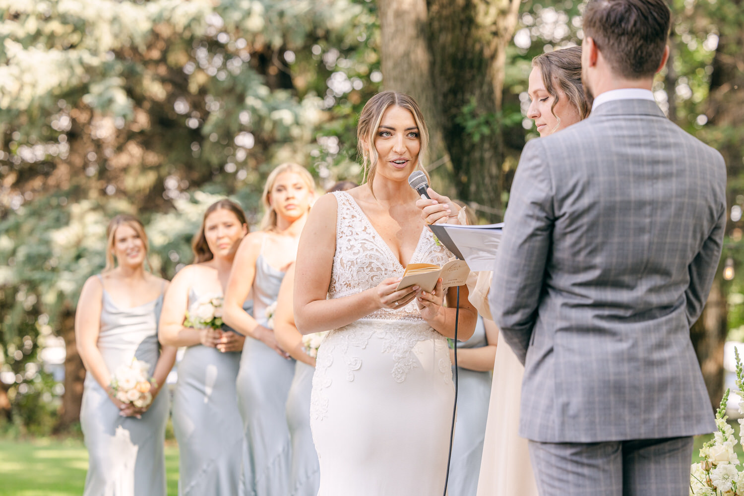 Bride speaking during the wedding ceremony with bridesmaids in background, surrounded by greenery.