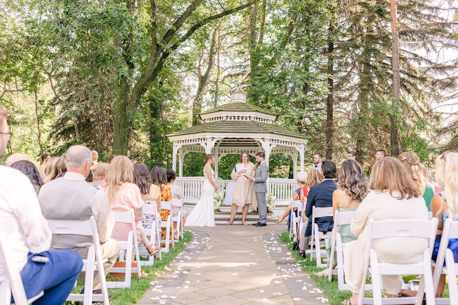 An outdoor wedding ceremony taking place in front of a gazebo surrounded by guests and lush greenery.