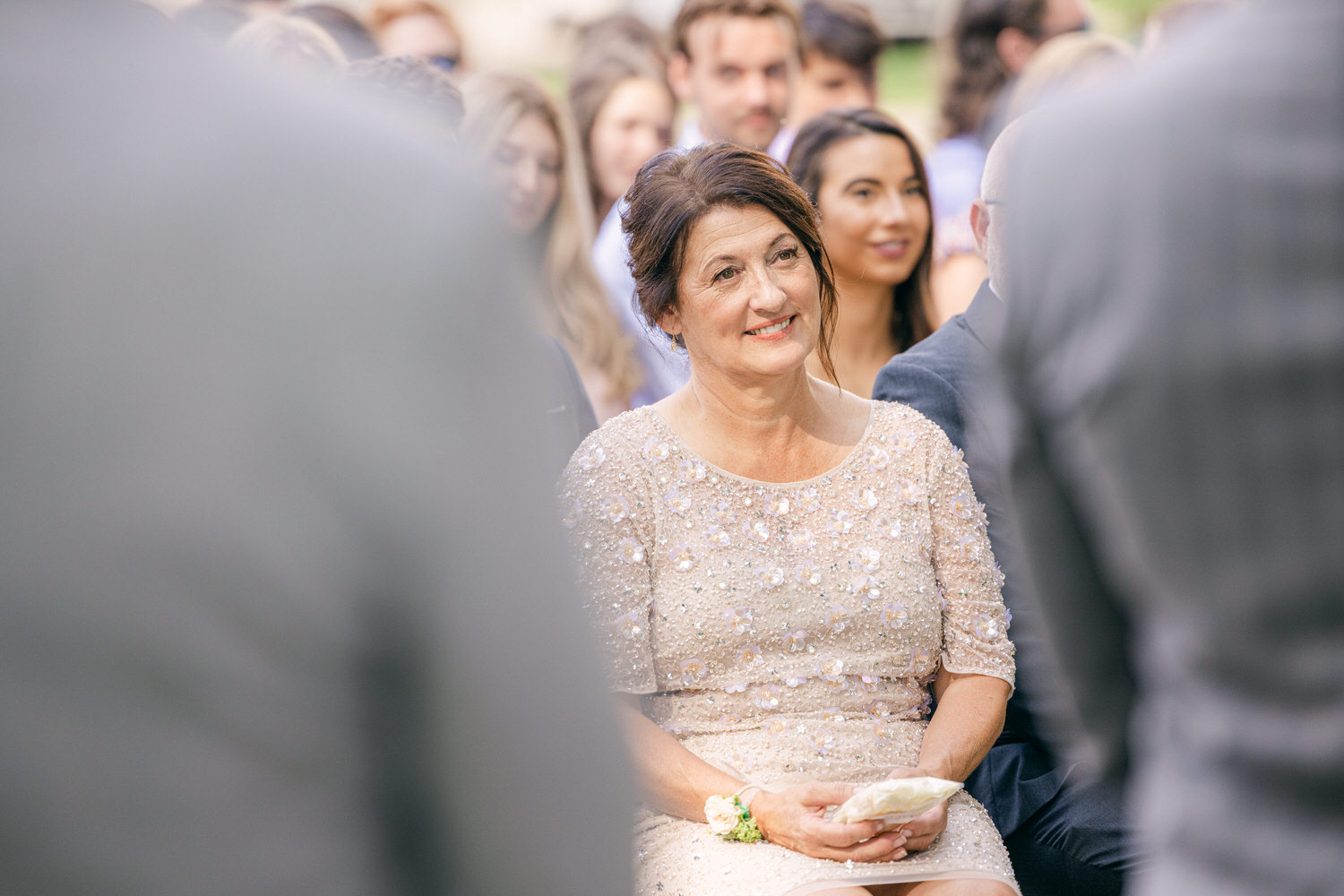 A smiling woman in a sequined dress sits among guests during a wedding ceremony, exuding joy and emotion.