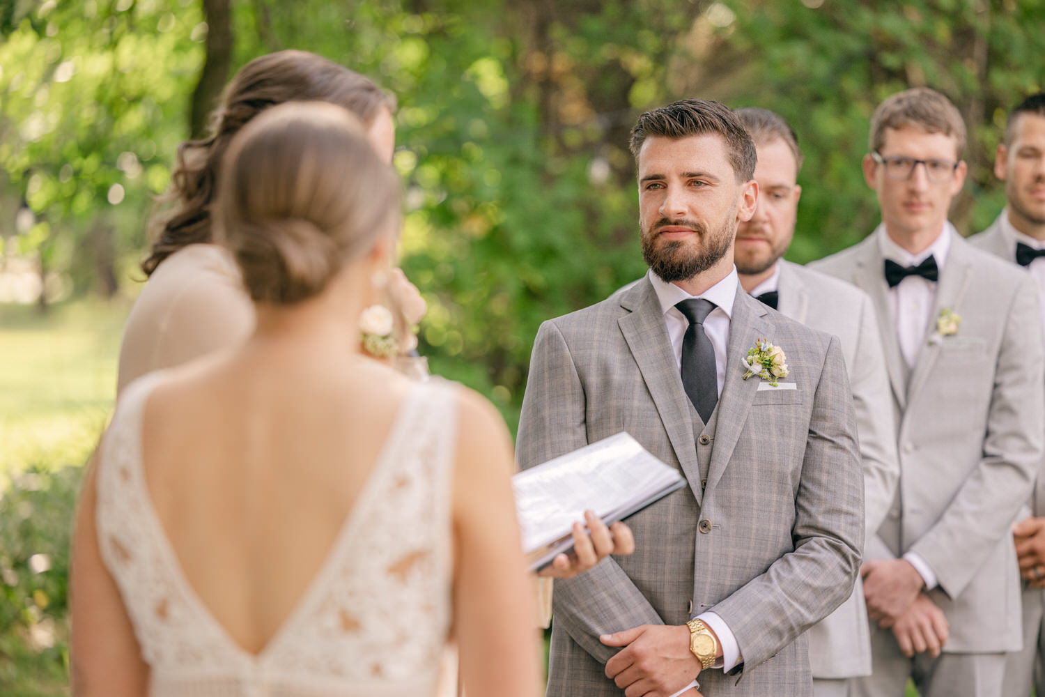 A groom stands attentively during an outdoor wedding ceremony while the officiant speaks, surrounded by groomsmen in light gray suits.