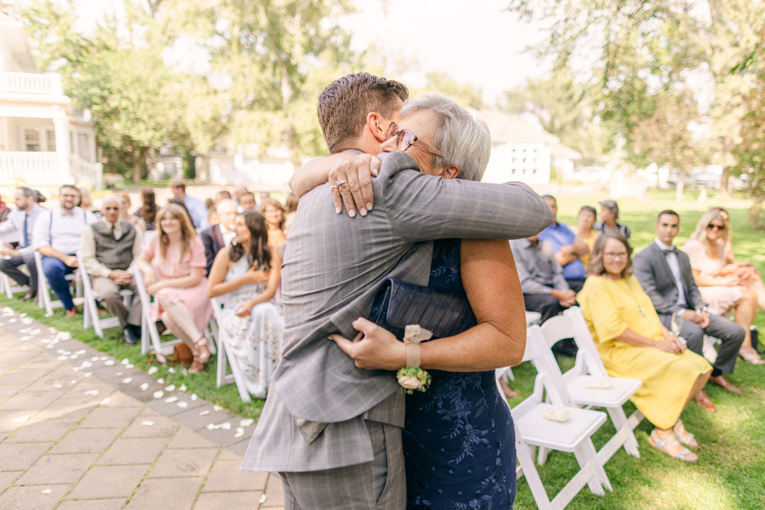 A joyful embrace between a man in a gray suit and a woman in a blue dress, surrounded by wedding guests seated in the background.