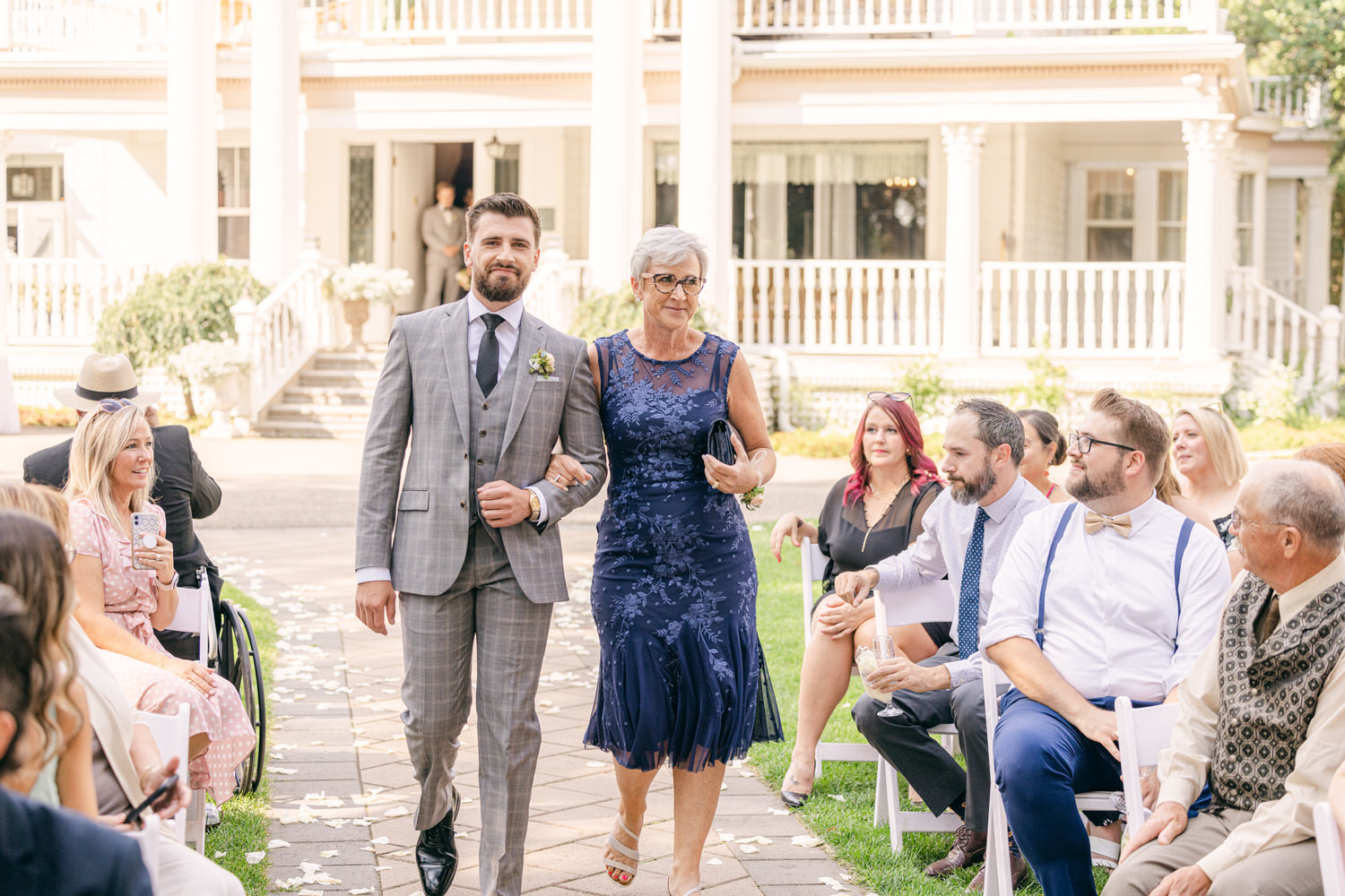 A groom in a gray plaid suit walks arm-in-arm with his mother, dressed in a blue floral gown, during a wedding ceremony. Guests watch attentively in a picturesque outdoor setting.