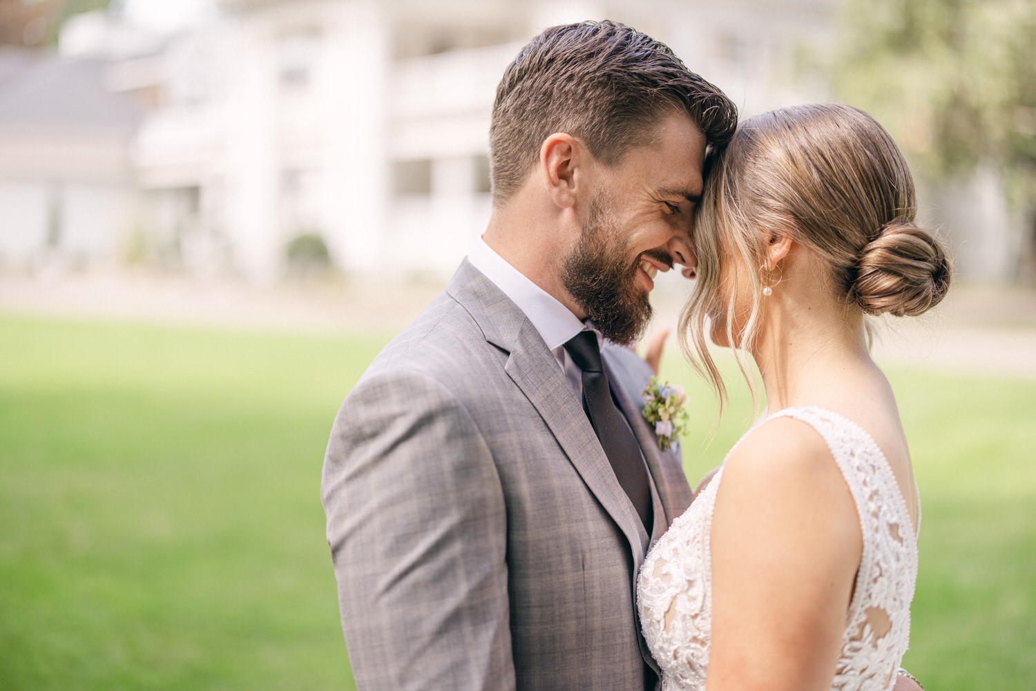 A couple sharing a tender moment, foreheads touching, surrounded by greenery and soft sunlight. The man in a light gray suit and the woman in a beautifully detailed wedding dress.