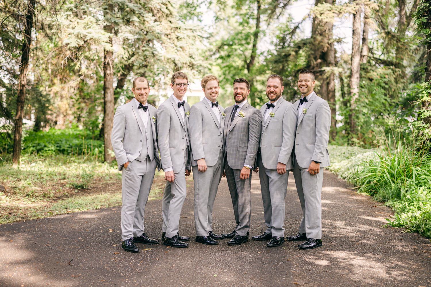 A group of six men dressed in gray suits and bow ties stand together outdoors, smiling in a lush green setting.